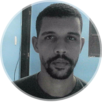 Foto de Marcos Marcelino com a feição de sério, usando camisa preta barba em um local fechado com parede azul, uma porta a direita dele na foto e uma janela ao fundo a esquerda dele
